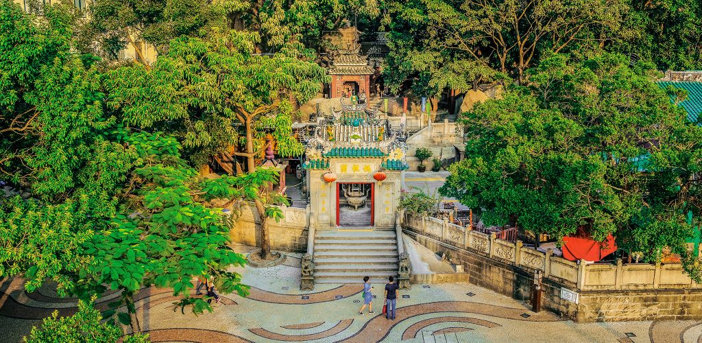 A colourful jungle temple in Macao with lush vegetation and some visitors strolling past.